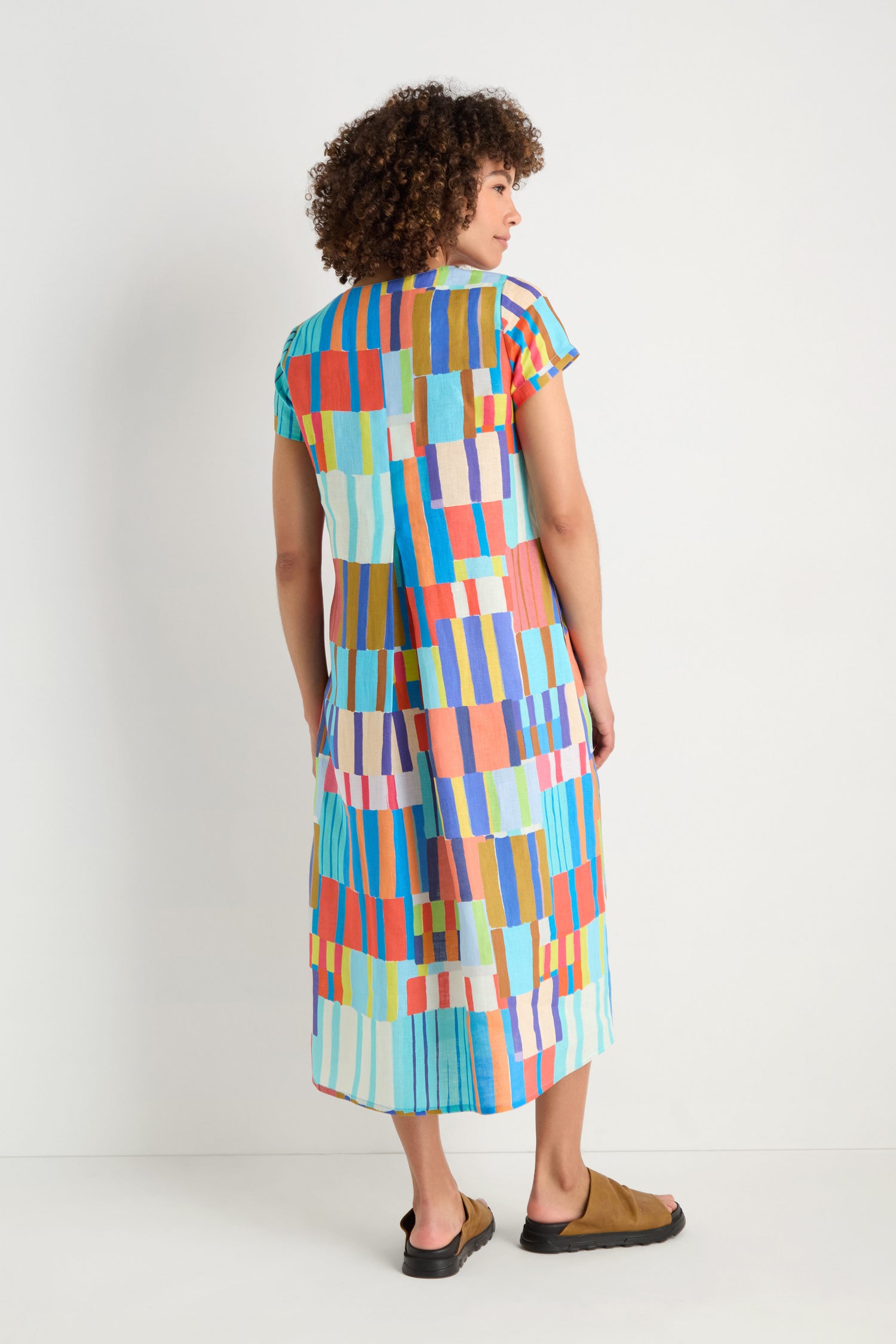 A person faces away, wearing the Painted Stripes A-Line Dress with colorful geometric patterns and brown slip-on sandals against a plain white background.