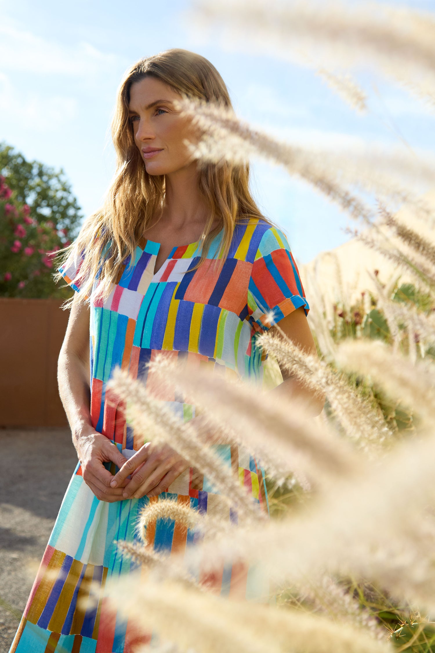 A woman in a colorful, geometric-patterned Painted Stripes A-Line Dress stands outdoors near tall grass, sunlight illuminating the scene.