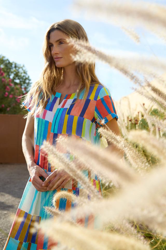 A woman in a colorful, geometric-patterned Painted Stripes A-Line Dress stands outdoors near tall grass, sunlight illuminating the scene.