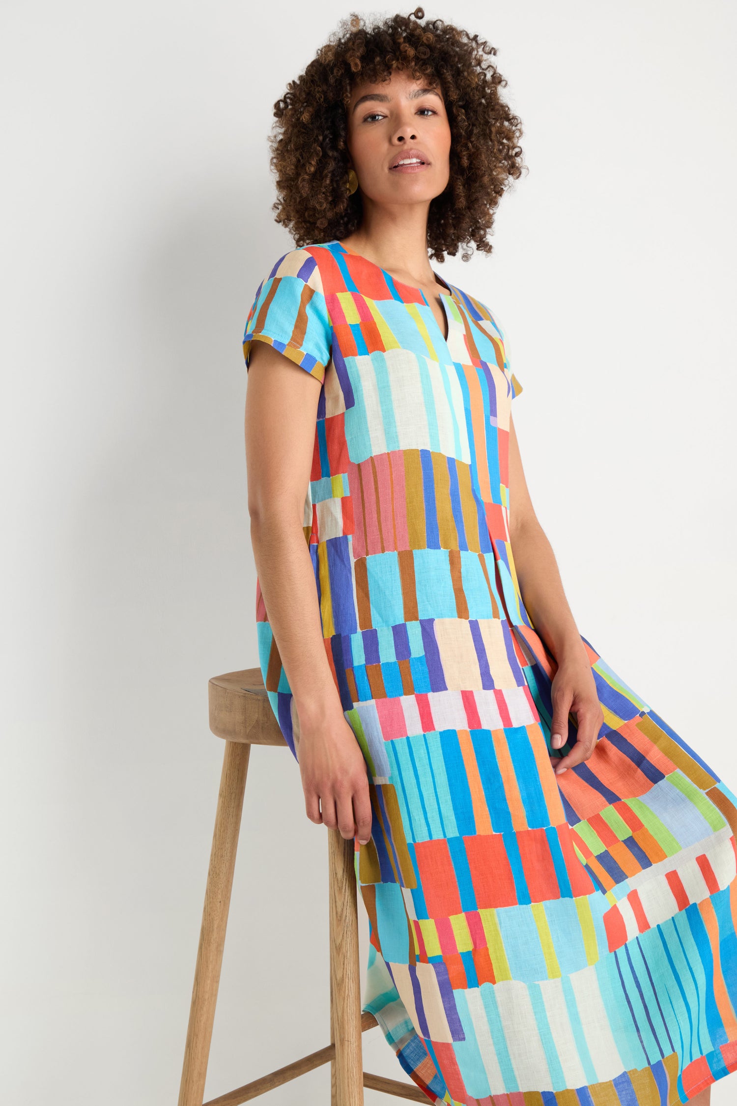 A woman with curly hair sits on a wooden stool wearing the Painted Stripes A-Line Dress, featuring colorful hand-painted geometric patterns, against a plain white background.