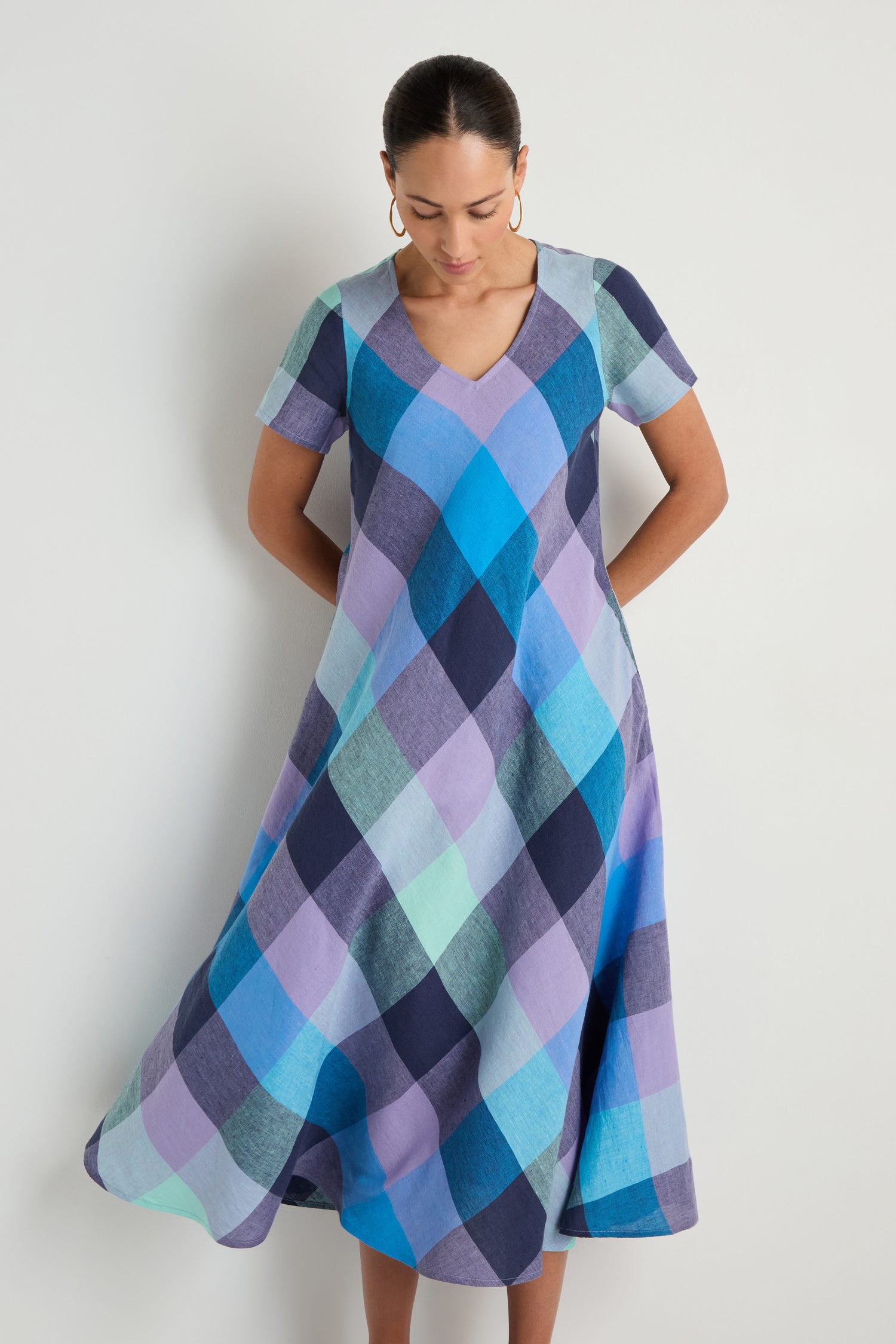 A person wears the Cross Dye Check Linen Maxi Dress, a short-sleeve V-neck maxi in blue, purple, and teal checkered linen, standing against a plain white background.