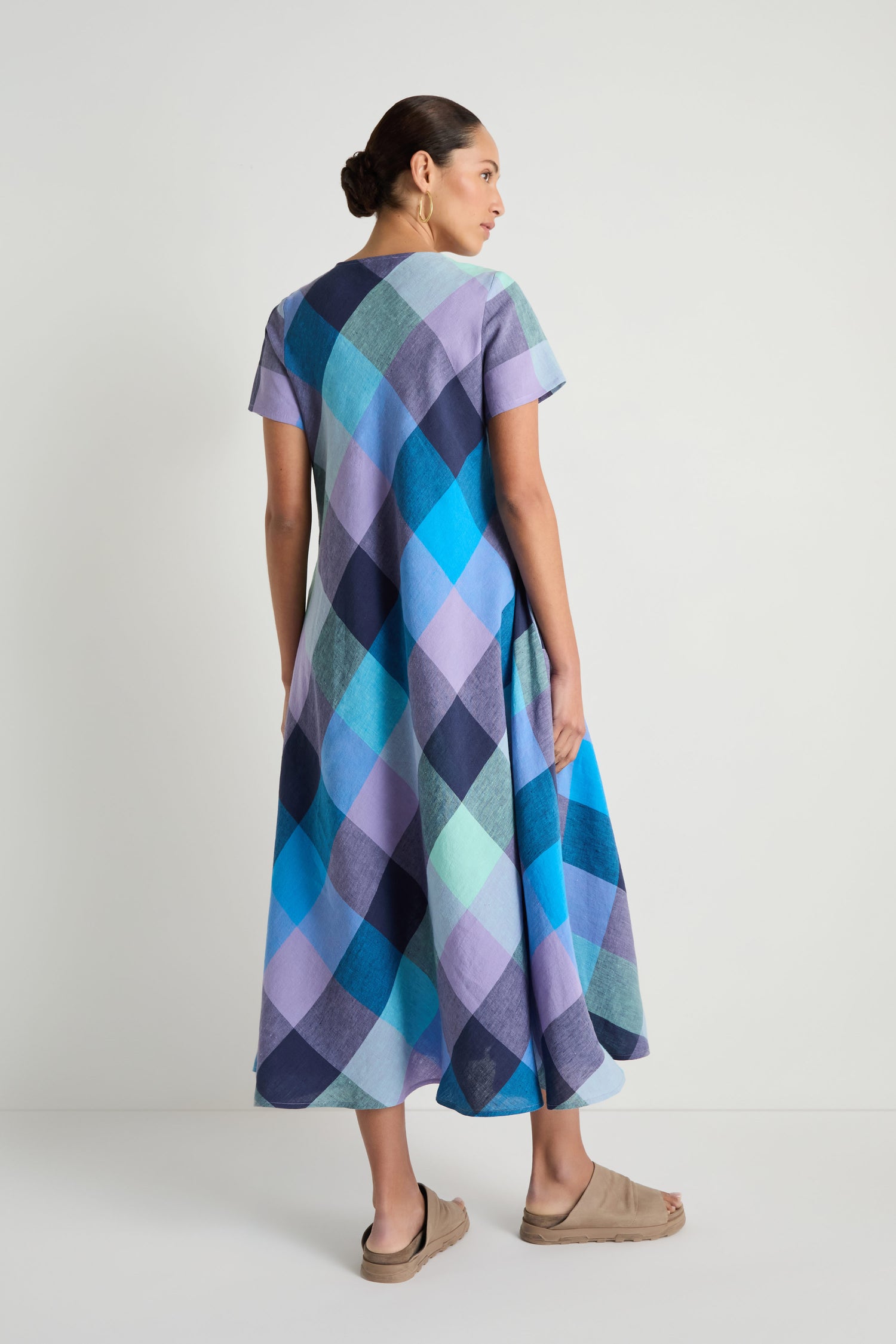 A woman faces away, wearing the Cross Dye Check Linen Maxi Dress in blue and purple tones with beige sandals, set against a plain white background.