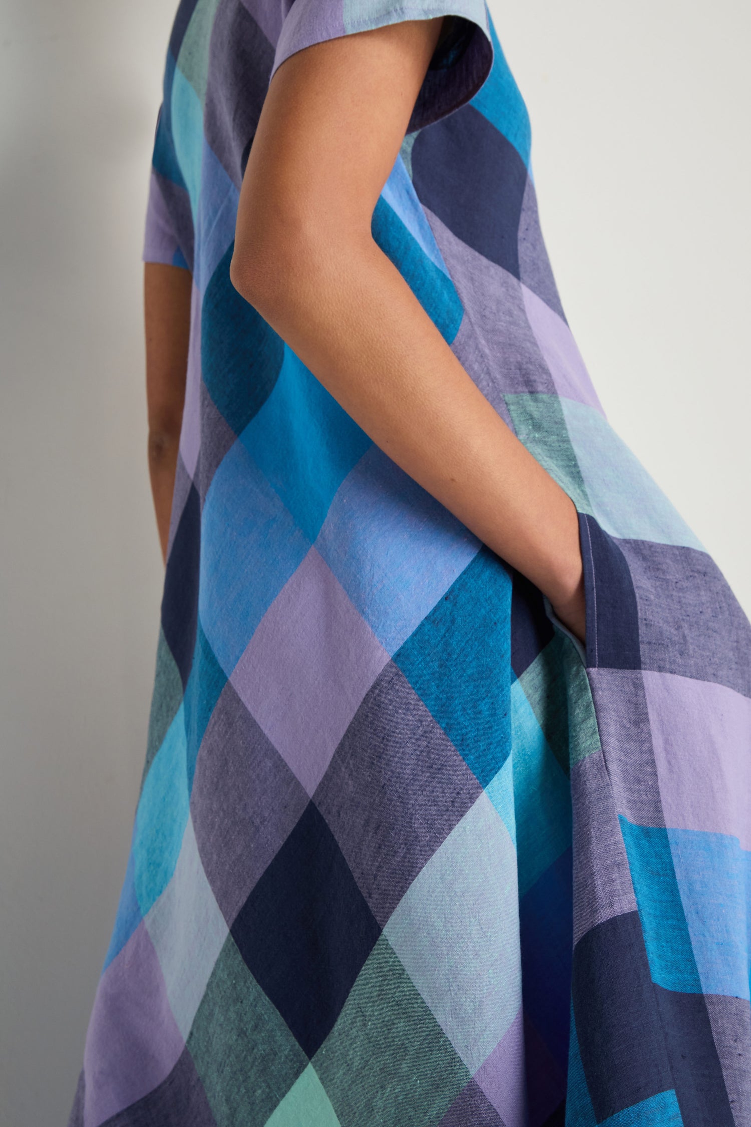 A person stands against a plain background with one hand in their pocket, wearing the Cross Dye Check Linen Maxi Dress—featuring short sleeves and large blue, purple, and teal check patterns.