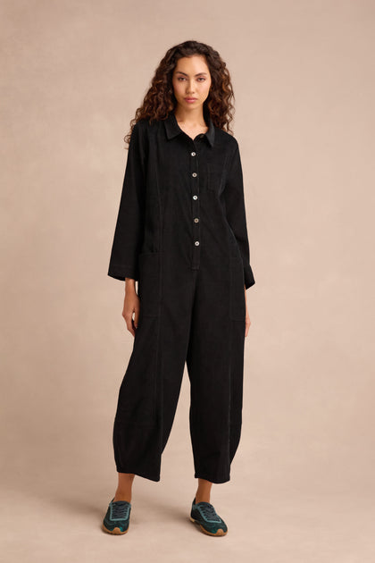 A person stands against a beige background wearing the Pinwale Cord Jumpsuit—crafted in pure cotton black cord with long sleeves and bubble leg details—paired with teal sneakers.