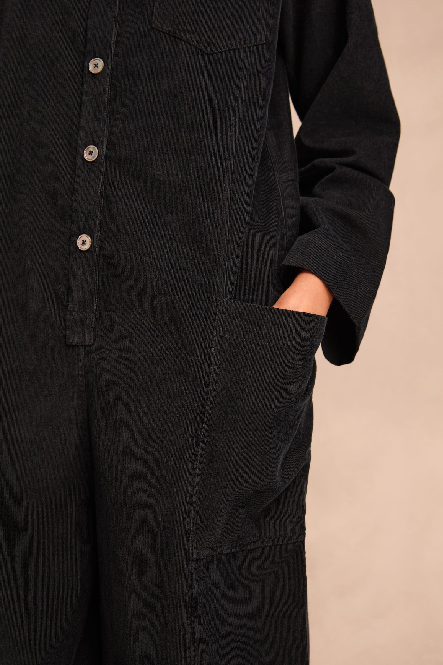 Close-up of a person wearing the Pinwale Cord Jumpsuit in black pure cotton, featuring buttons and a bubble leg design, standing with one hand in a front pocket against a neutral background.