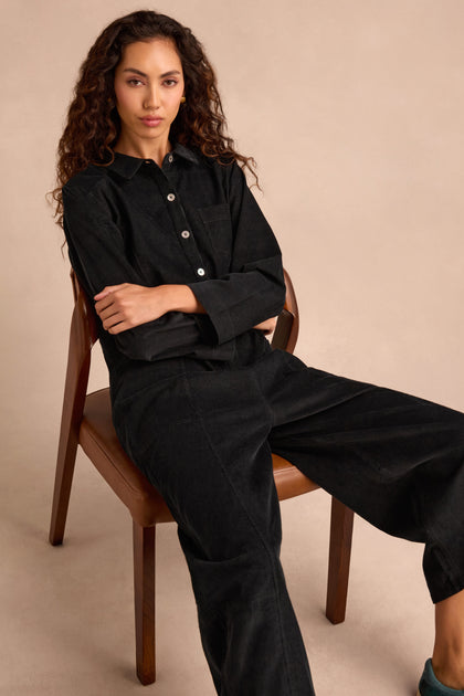 A person with long curly hair sits on a wooden chair, wearing the Pinwale Cord Jumpsuit—black, pure cotton, with bubble leg detail—arms crossed and a neutral expression, against a beige background.