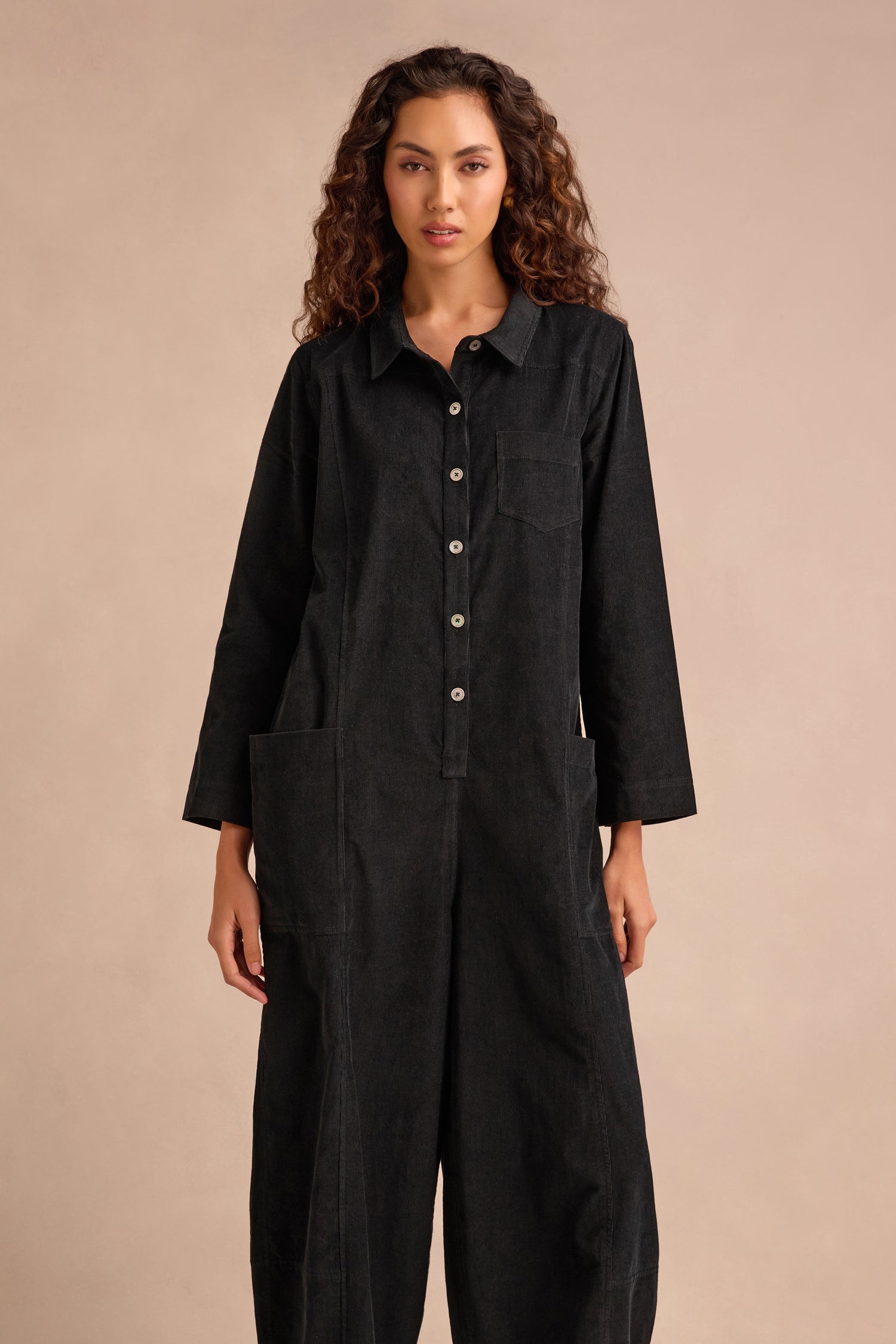 A person with long curly hair wearing the Pinwale Cord Jumpsuit— a black, pure cotton button-up style with long sleeves and bubble legs—stands against a plain beige background.