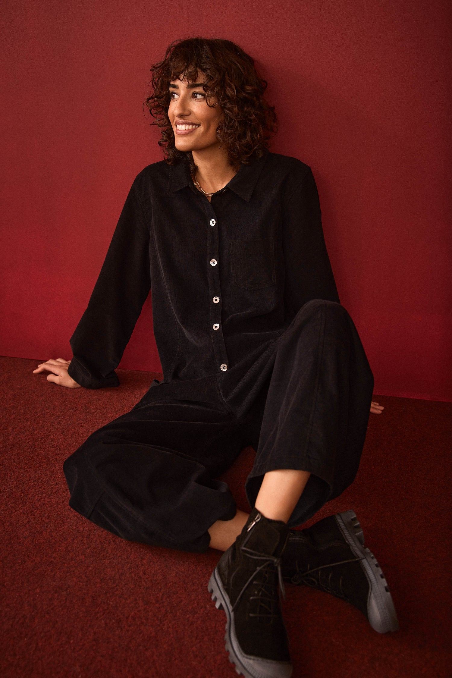 A person with curly hair sits on a red floor against a red wall, smiling and looking slightly to the side while wearing the Pinwale Cord Jumpsuit and boots.