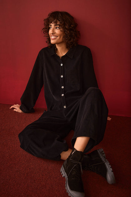 A person with curly hair sits on a red floor against a red wall, smiling and looking slightly to the side while wearing the Pinwale Cord Jumpsuit and boots.