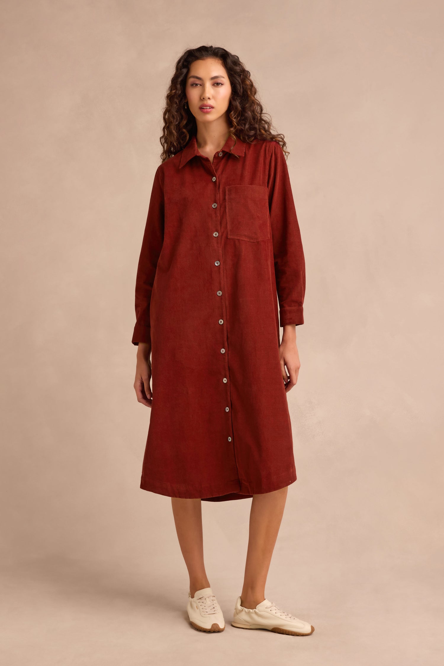 A woman wears the Conker Pinwale Cord Shirt Dress, a rust-colored, long-sleeve, button-down cord shirt dress featuring a chest pocket. She stands on a neutral background paired with white sneakers.