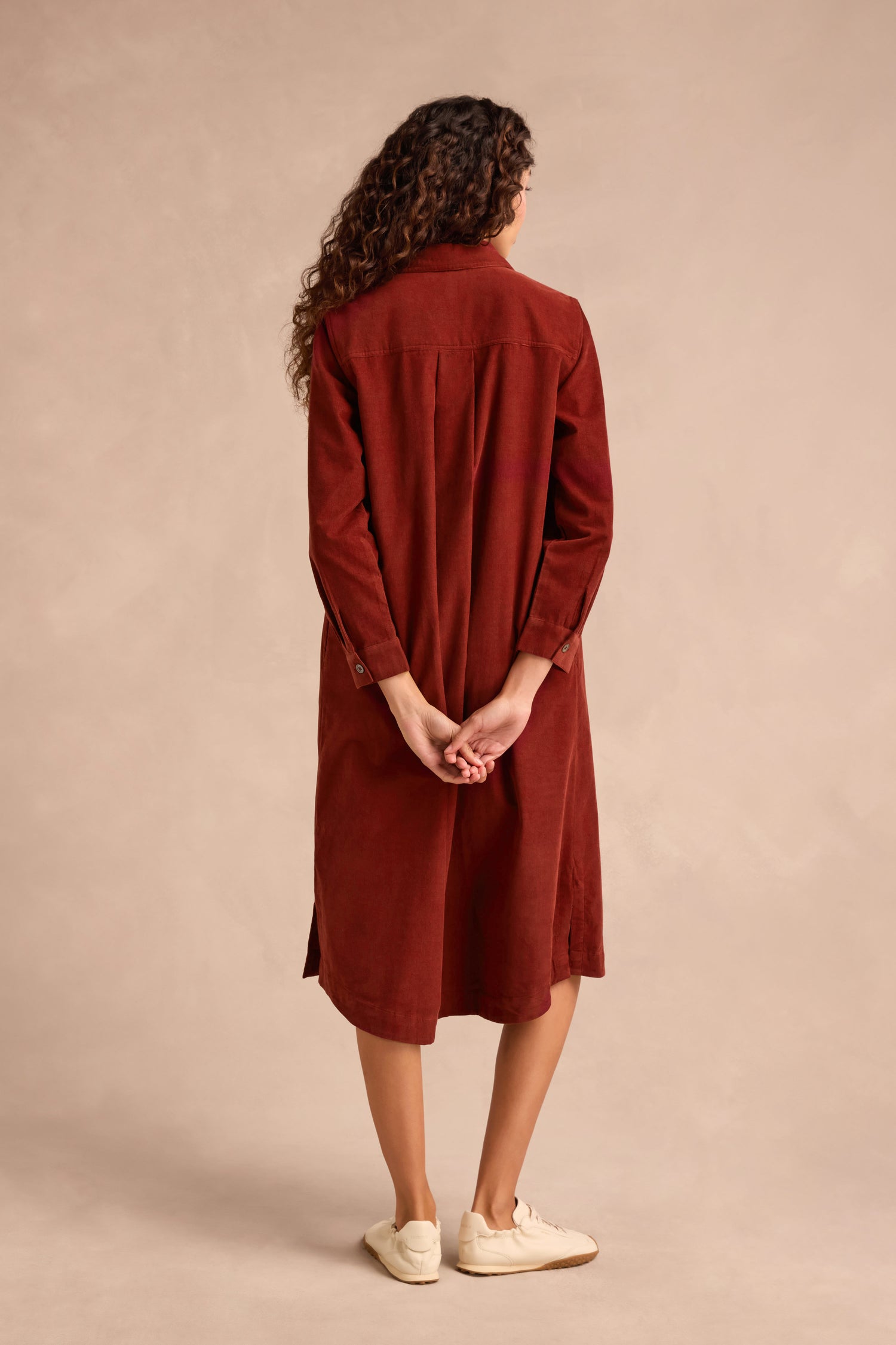 A woman with curly hair stands facing away, wearing the Conker Pinwale Cord Shirt Dress—a long-sleeved, rust-colored cotton cord dress—and cream sneakers, with her hands clasped behind her back.