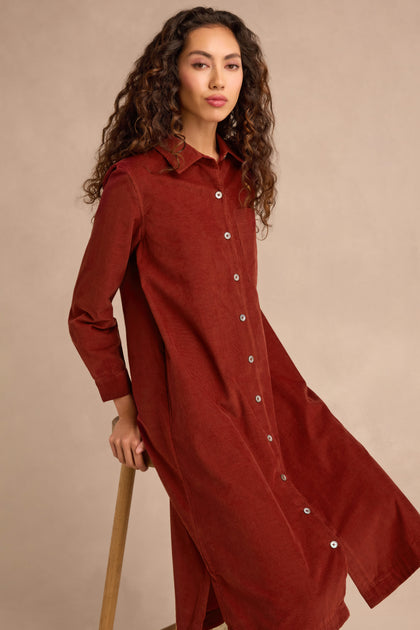A woman wearing the Conker Pinwale Cord Shirt Dress, a long-sleeve, button-down rust-colored cotton cord dress, sits on a wooden stool against a beige background.