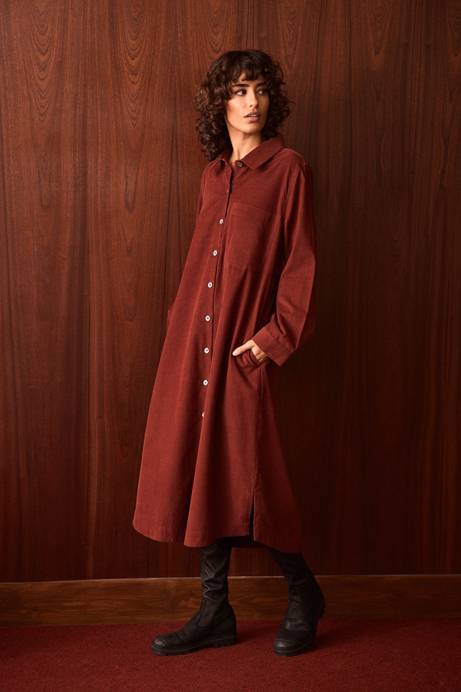 A person stands against a wood-paneled wall wearing the Conker Pinwale Cord Shirt Dress and black boots.