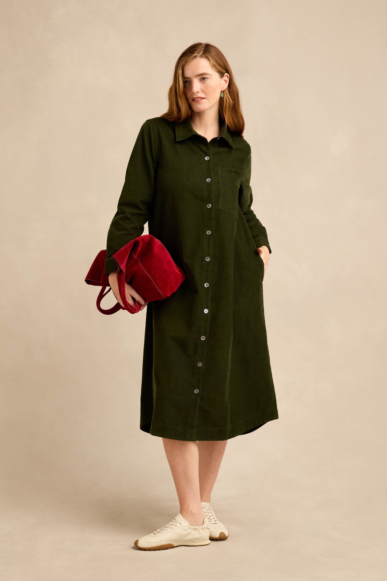 A woman wearing the Forest Pinwale Cord Shirt Dress, holding a red handbag and standing in white sneakers against a beige background.