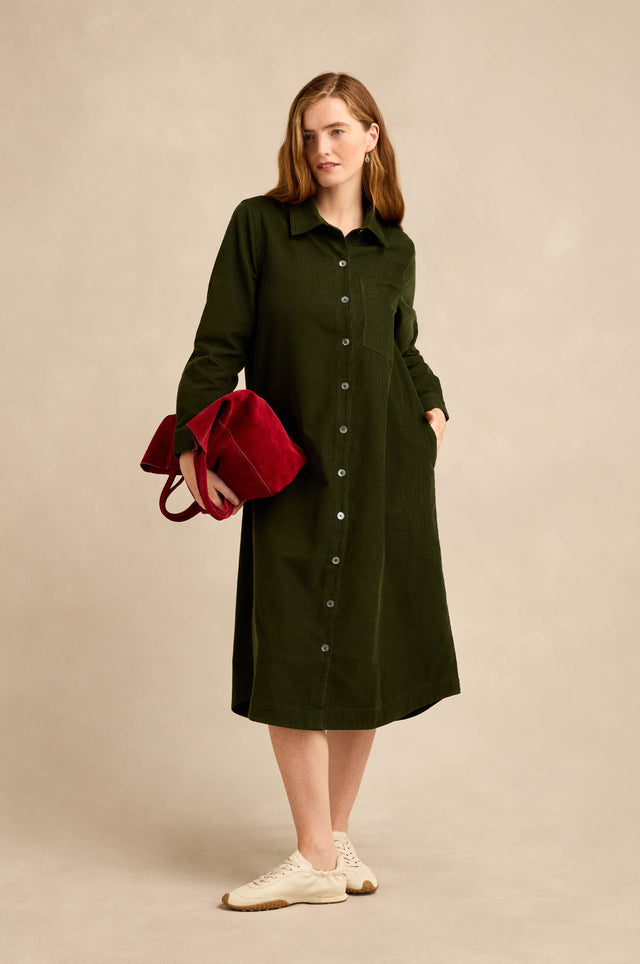A woman wearing the Forest Pinwale Cord Shirt Dress, holding a red handbag and standing in white sneakers against a beige background.