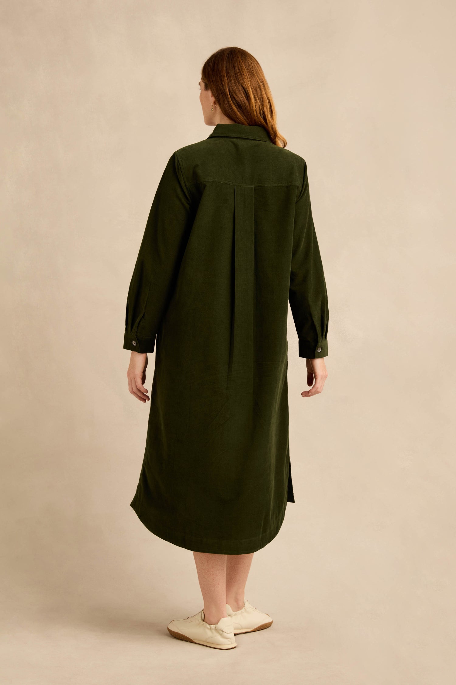 A person with long hair stands facing away, wearing the Forest Pinwale Cord Shirt Dress—a knee-length, long-sleeve green piece—paired with white sneakers against a plain beige background.