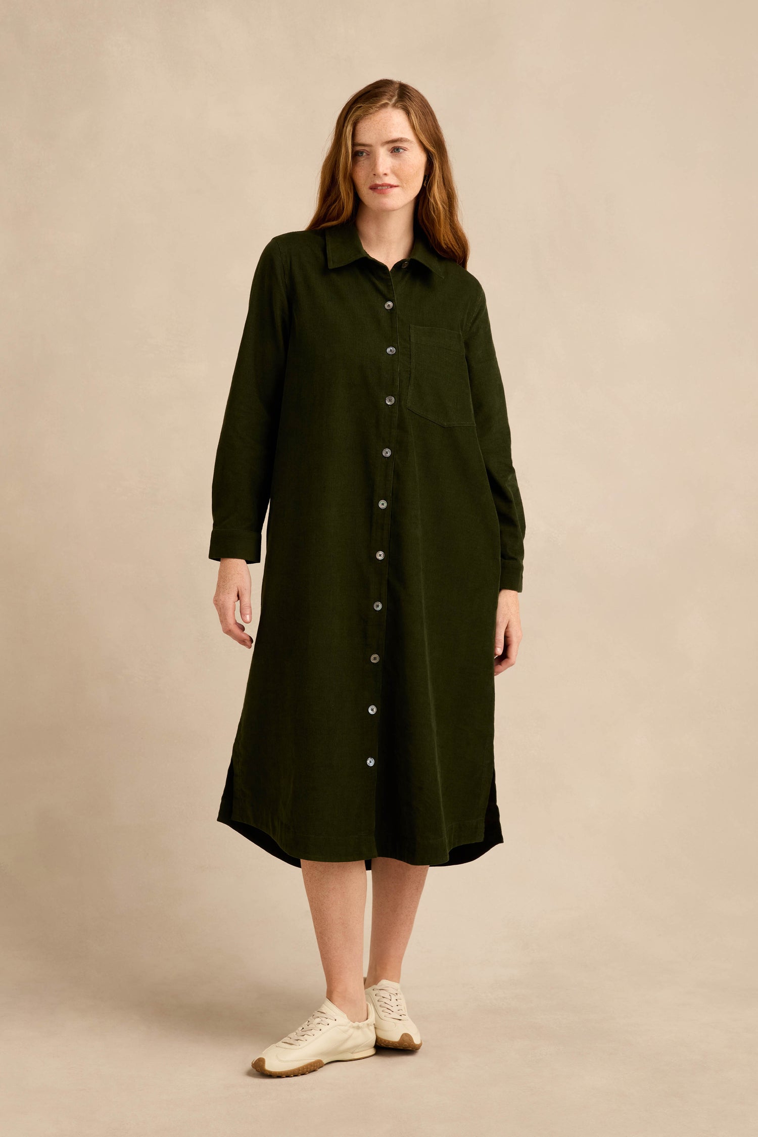 A woman stands against a beige background, wearing the Forest Pinwale Cord Shirt Dress—a long-sleeved, button-down green midi dress crafted from soft cotton cord. She completes her casual yet refined look with light-colored sneakers.