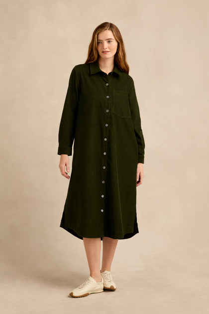A woman stands against a beige background, wearing the Forest Pinwale Cord Shirt Dress—a long-sleeved, button-down green midi dress crafted from soft cotton cord. She completes her casual yet refined look with light-colored sneakers.
