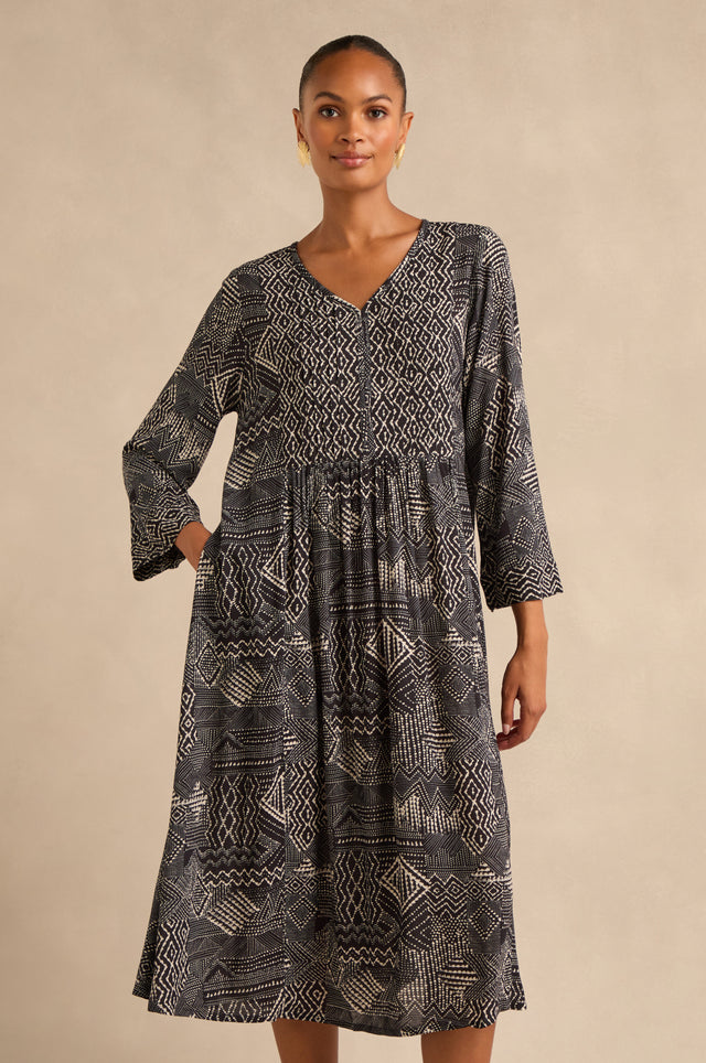 A woman stands against a neutral background, wearing the Mono Patchwork Ikat Dress—an A-line silhouette with long sleeves, a V-neckline, a gathered waist, and a striking black and white pattern.