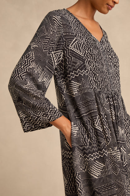 A person is photographed from the side, wearing the Mono Patchwork Ikat Dress—a long-sleeved, black and white geometric patterned design with an A-line silhouette and a pocket where one hand rests.