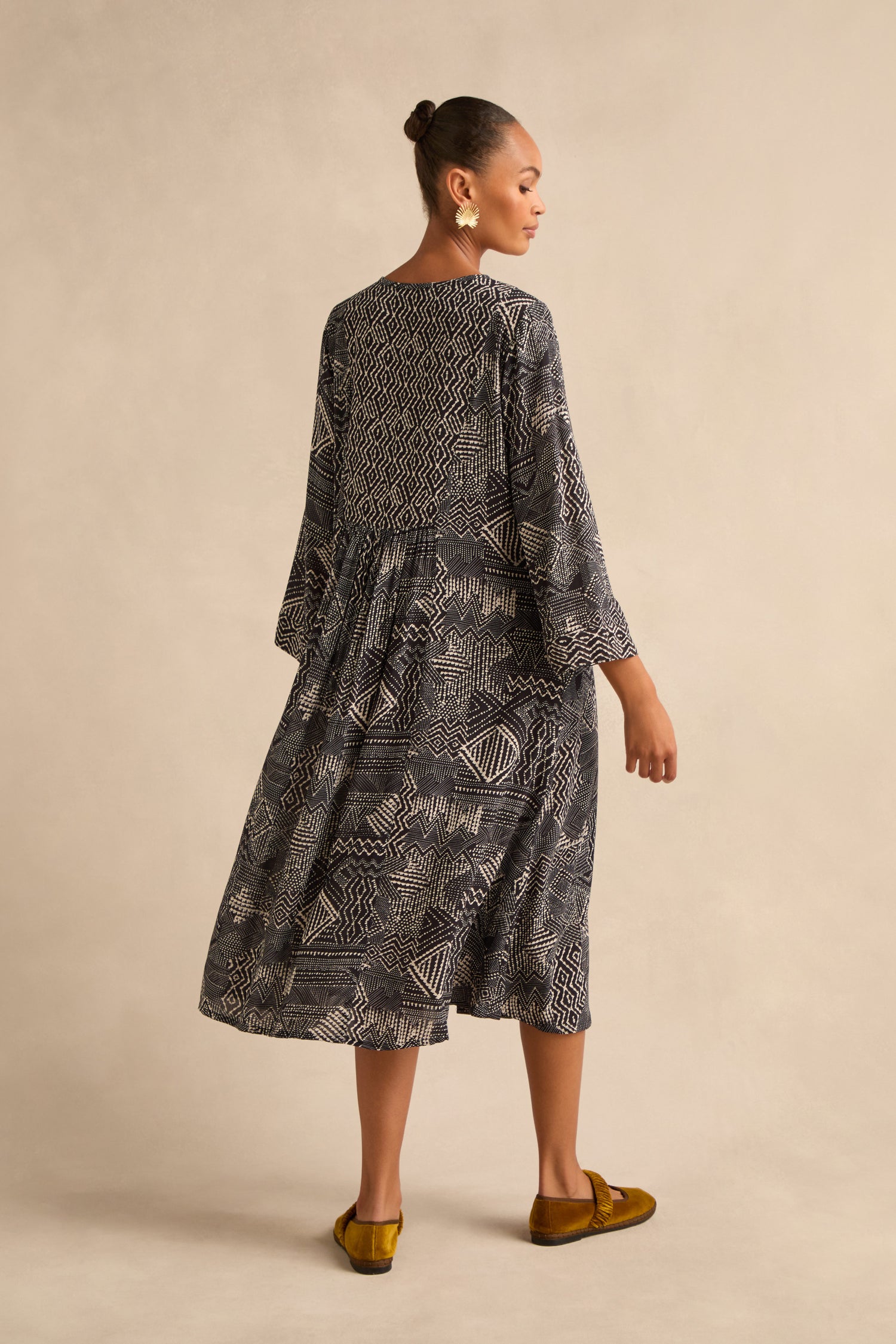 A woman stands sideways against a neutral beige background, wearing the Mono Patchwork Ikat Dress—featuring an A-line silhouette and long sleeves—paired with brown flats.