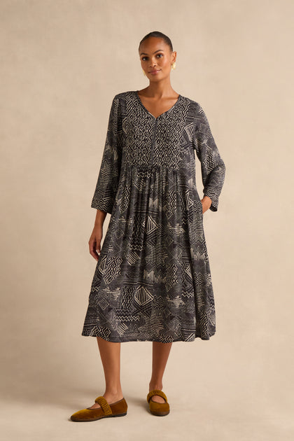 A woman stands against a beige background wearing the Mono Patchwork Ikat Dress, an ankle-length, long-sleeved A-line dress featuring white geometric patterns, paired with brown flat shoes.