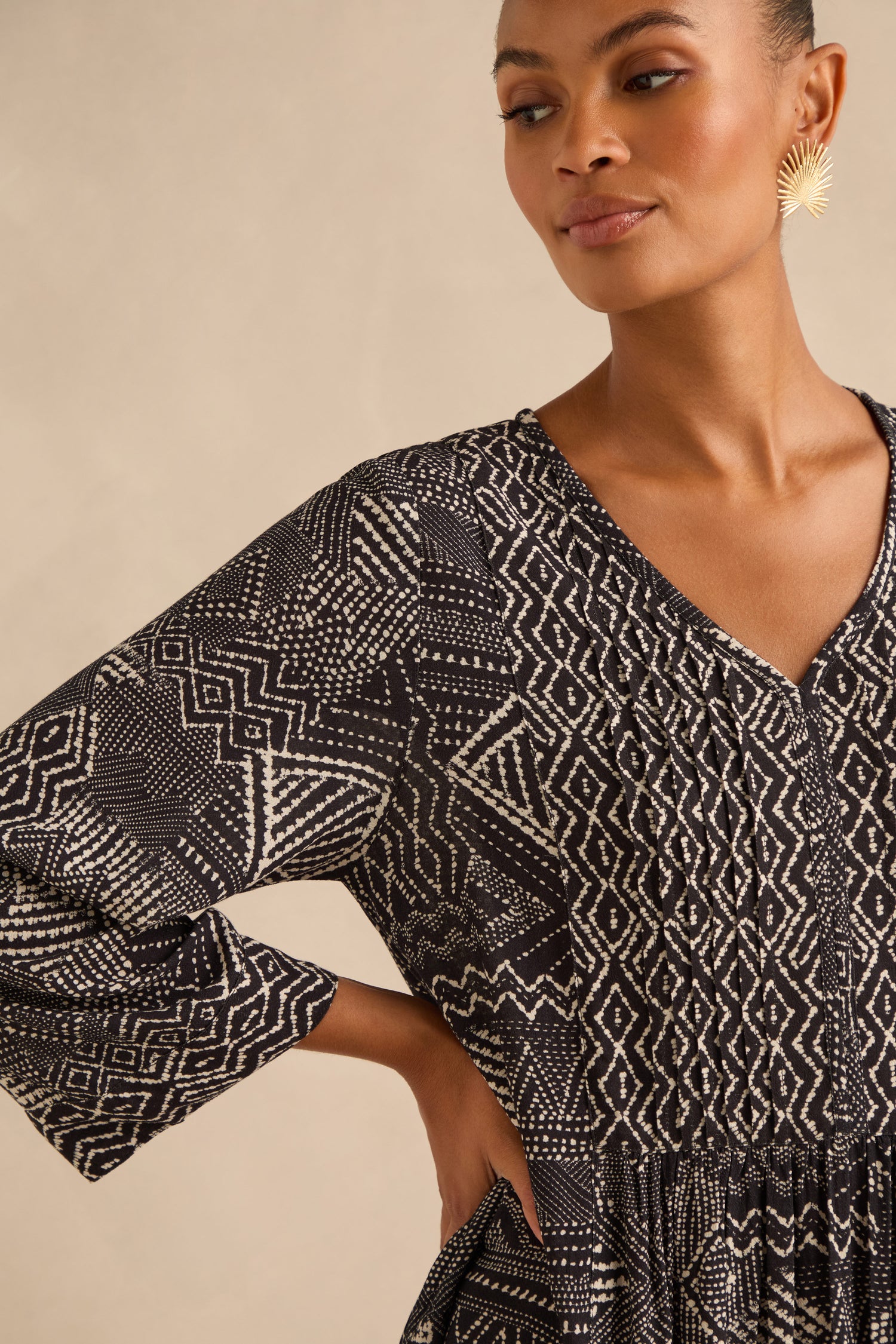 A woman wearing the Mono Patchwork Ikat Dress—a long-sleeve, black and white geometric A-line dress—paired with gold sunburst earrings and her hair pulled back, stands against a neutral background.