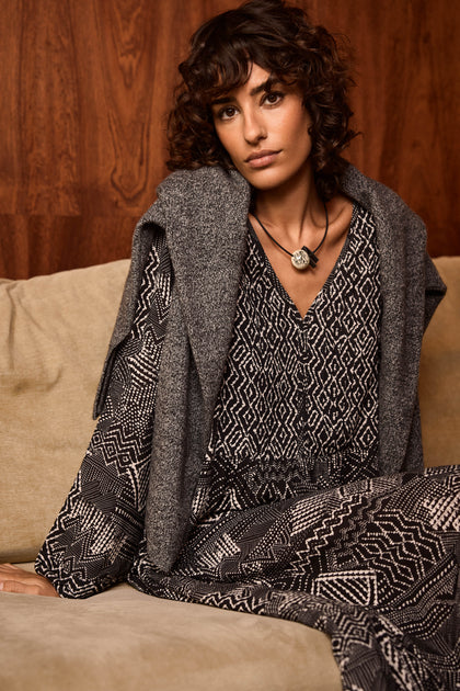 A woman with curly hair sits on a beige sofa, wearing the Monochrome Batik Print Dress in an A-line silhouette, a gray sweater draped over her shoulders, and a pendant necklace, with a wooden wall as the background.