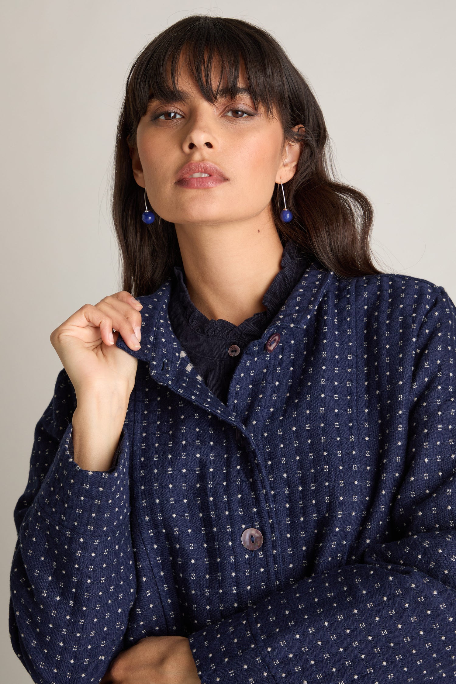 A person with long dark hair and bangs, wearing blue earrings and an Indigo Stitch Wool Cotton Jacket from the product line, stands against a light background, exuding an Autumnal style.