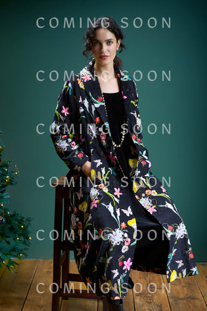 A person sits on a stool wearing a Velvet Embellished Vintage Floral Coat over a black outfit, with a decorated Christmas tree partially visible on the left.