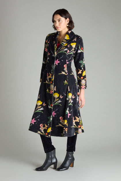 A person wearing the Velvet Vintage Floral Coat, which features a fit-and-flare silhouette, pairs it with black pants and boots while standing against a plain gray background.