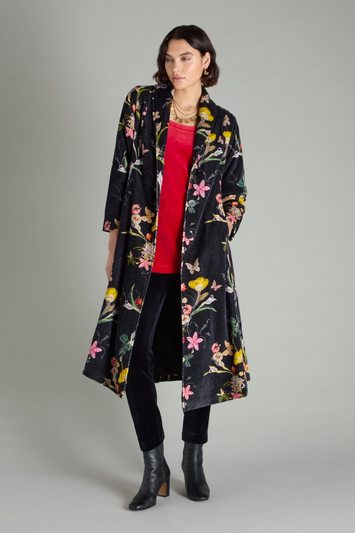 An individual dressed in a Velvet Vintage Floral Coat featuring a shawl collar, paired with a red top, black pants, and black boots, is positioned against a gray backdrop.