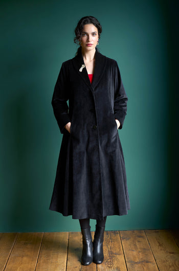 A person stands against a teal background wearing the Cotton Velvet Print Lined Coat, characterized by its long black design and shawl collar, paired with black boots and hands in pockets.