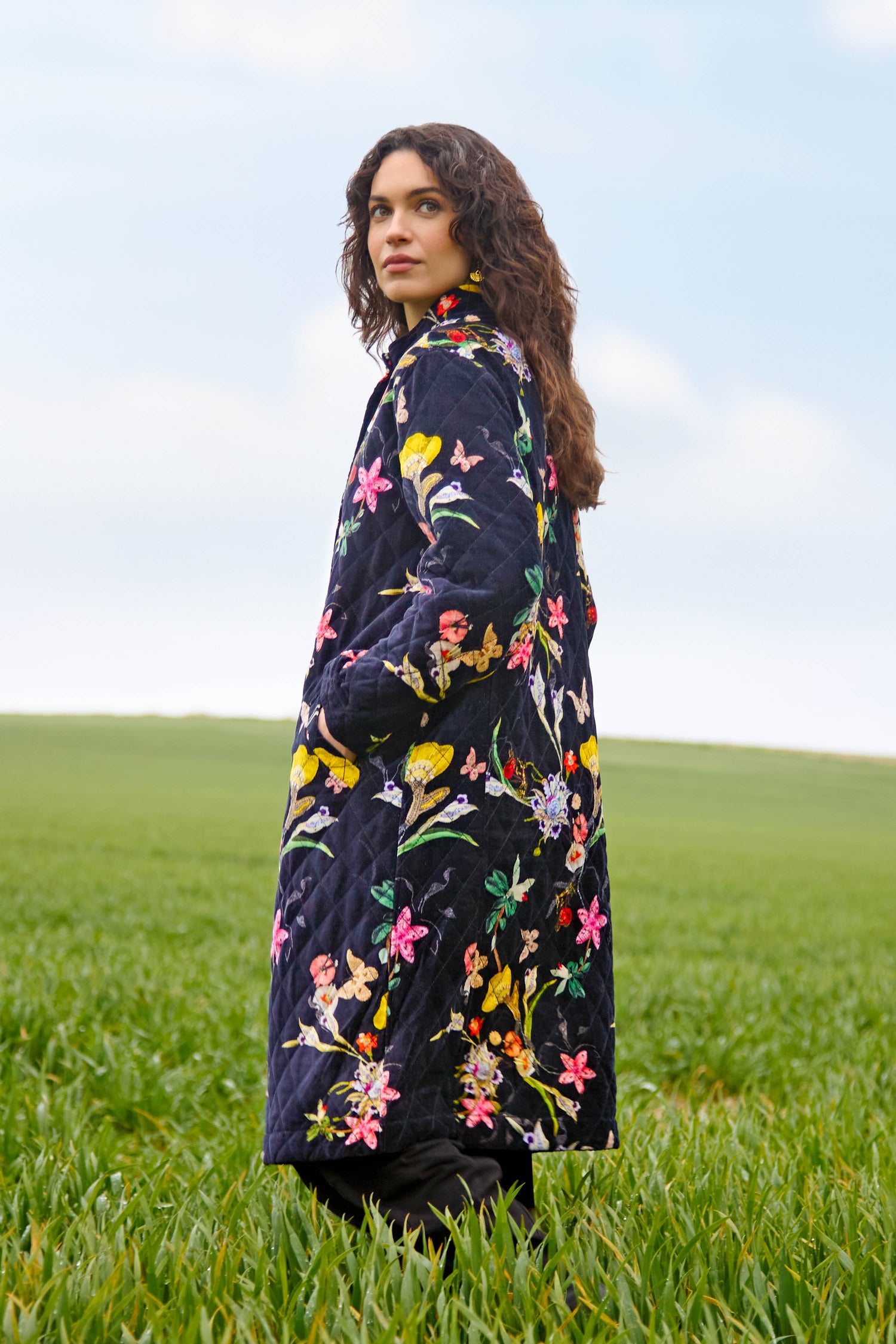 A person stands in a grassy field, wearing the Velvet Vintage Floral Quilted Coat with colorful floral embroidery and subtle diamond quilted padding, looking over their shoulder against a cloudy sky.