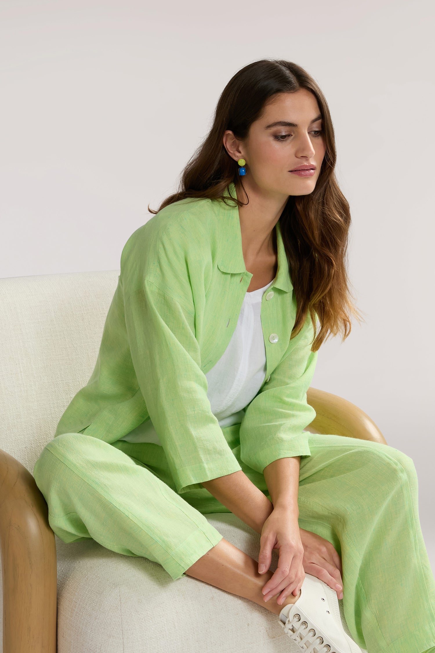 A woman wearing a light green outfit with a Linen Ticking Stripe Boxy Jacket sits on a white chair with wooden arms, looking down. She has long brown hair and is wearing colorful earrings.