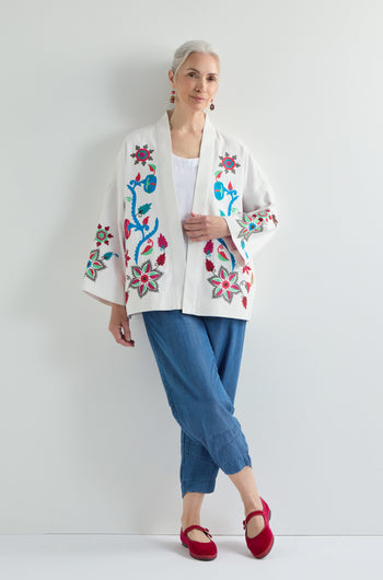 A woman with gray hair in a bun stands against a white wall, showcasing effortless Bohemian charm in the Boho Embroidery Jacket, styled with blue pants, a white top, and red shoes.
