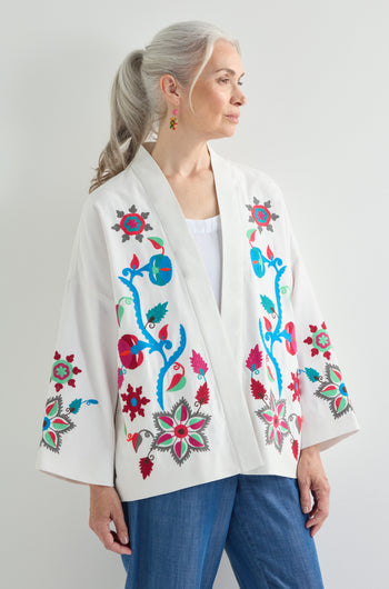 A woman with long gray hair in a ponytail wears the Boho Embroidery Jacket, featuring colorful floral patterns, along with blue jeans and floral earrings, standing against a plain background and exuding Bohemian charm.