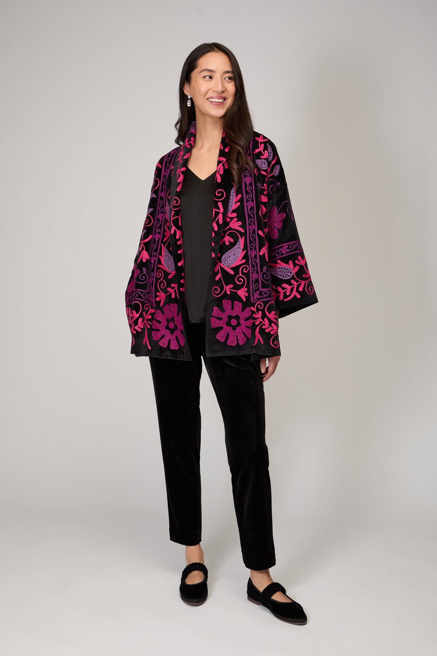 A woman stands smiling against a light background, wearing black pants and shoes paired with the Winter Embroidered Jacket—a loose black jacket featuring magenta embroidery, styled as a winter jacket.