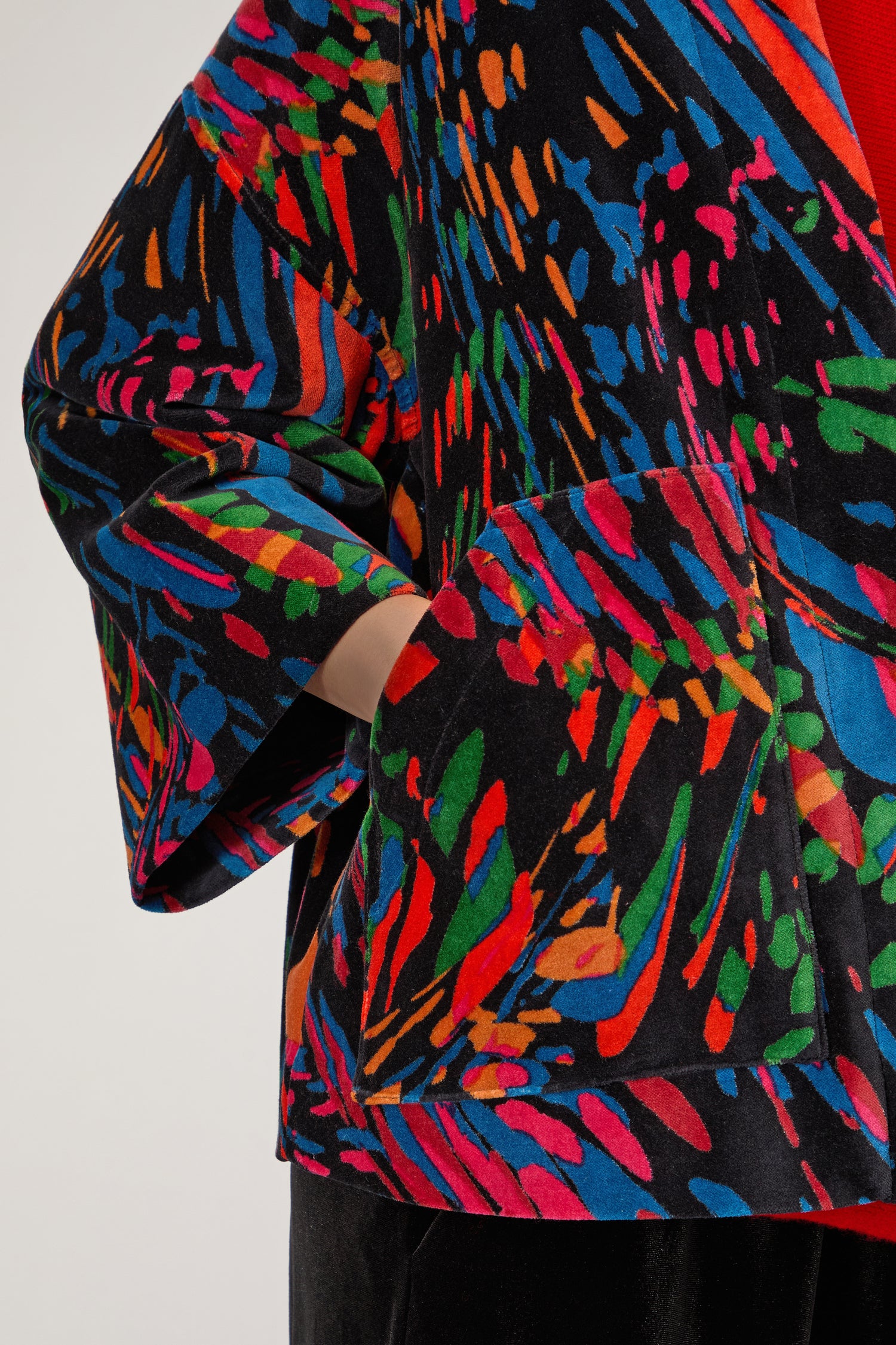 Wearing the Skyfire Velvet Velvet Jacket—a bold black piece with vivid abstract patterns—a person stands with one hand in a front pocket, making it perfect for standout evening looks.