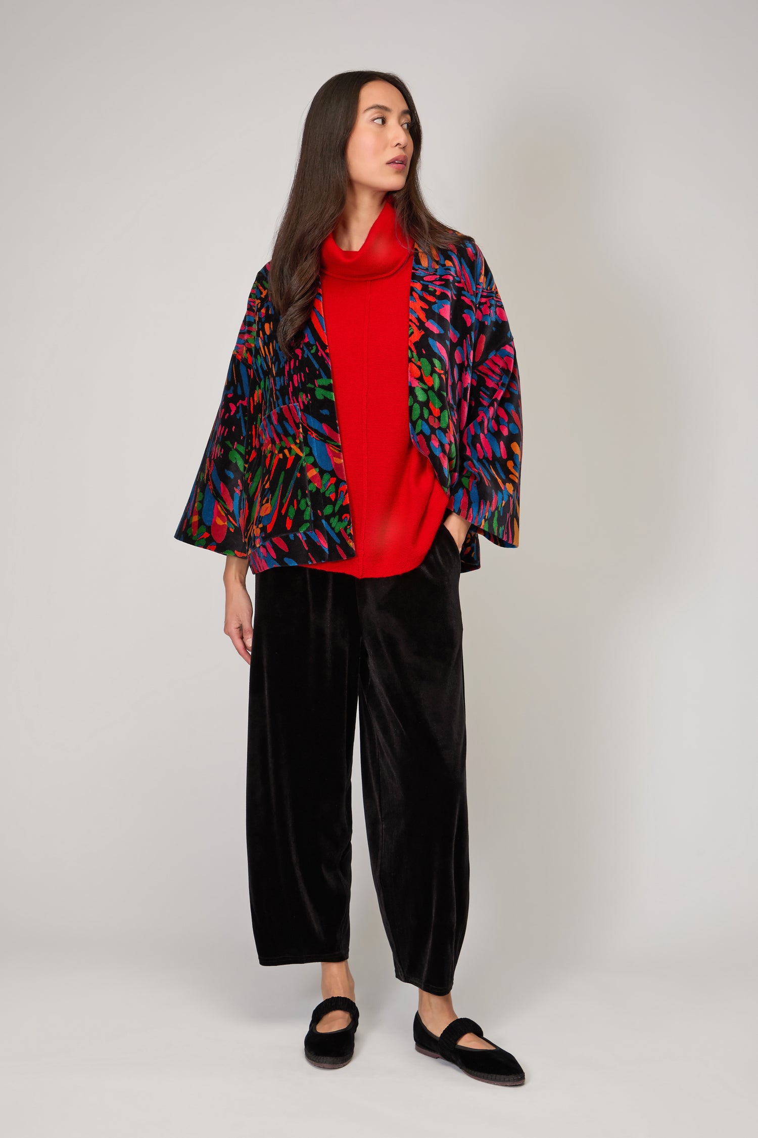 A woman stands against a plain background, wearing the Skyfire Velvet Velvet Jacket as a statement layer over a red turtleneck, paired with black wide-leg pants and black flats.