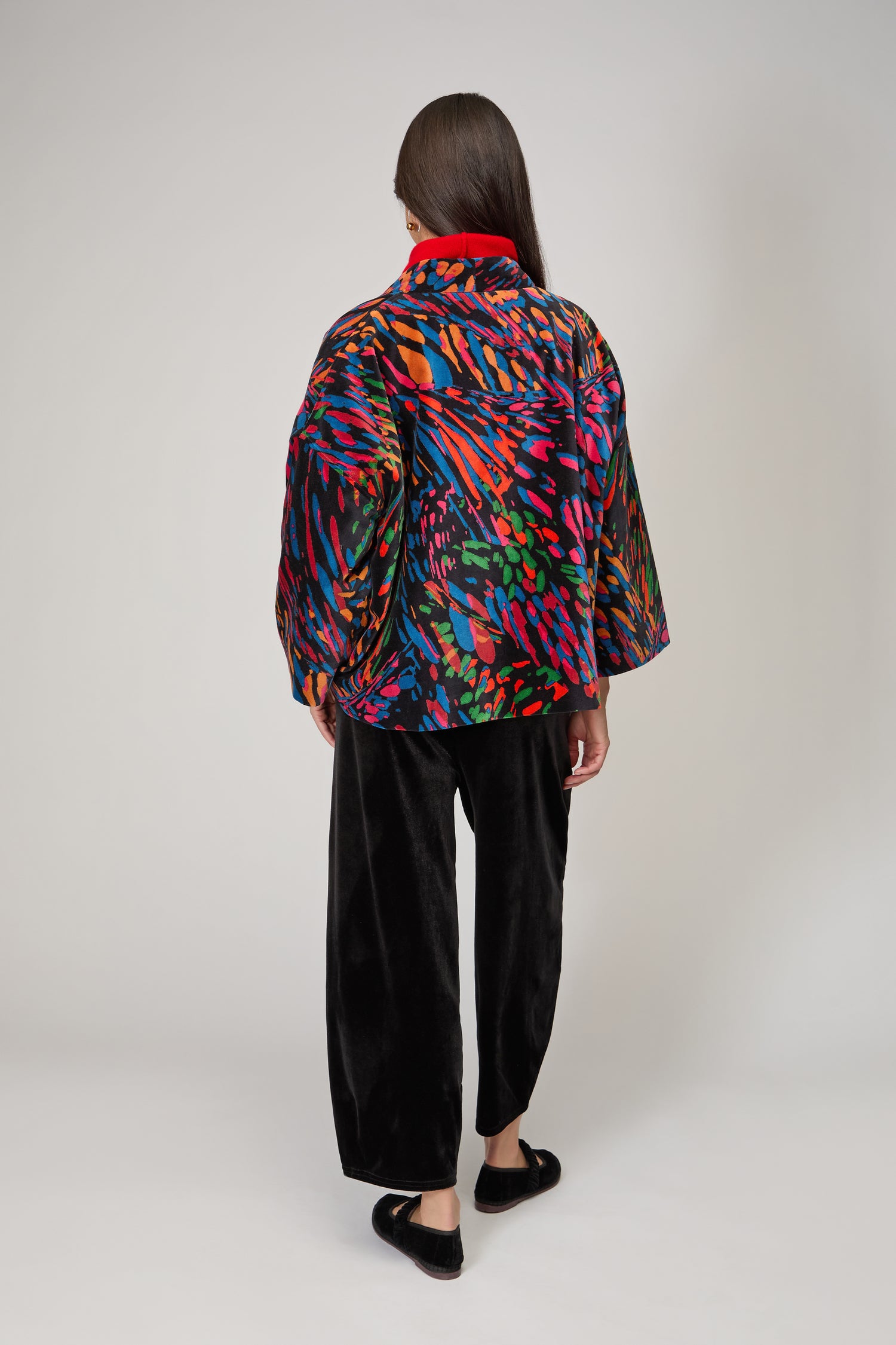 A person stands facing away against a plain light background, wearing the Skyfire Velvet Velvet Jacket—a colorful statement layer with wide sleeves—paired with black pants and black shoes.