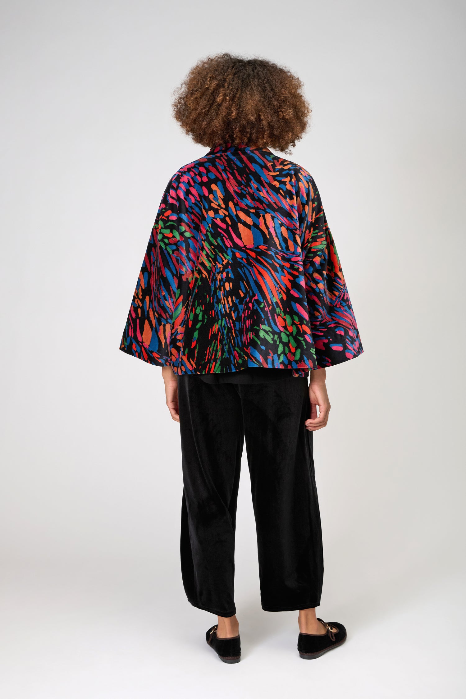 A person stands facing away, styled in the Skyfire Velvet Jacket—a loose, multicolored patterned piece with wide sleeves—paired with black pants and shoes against a plain background, perfect for standout evening looks.
