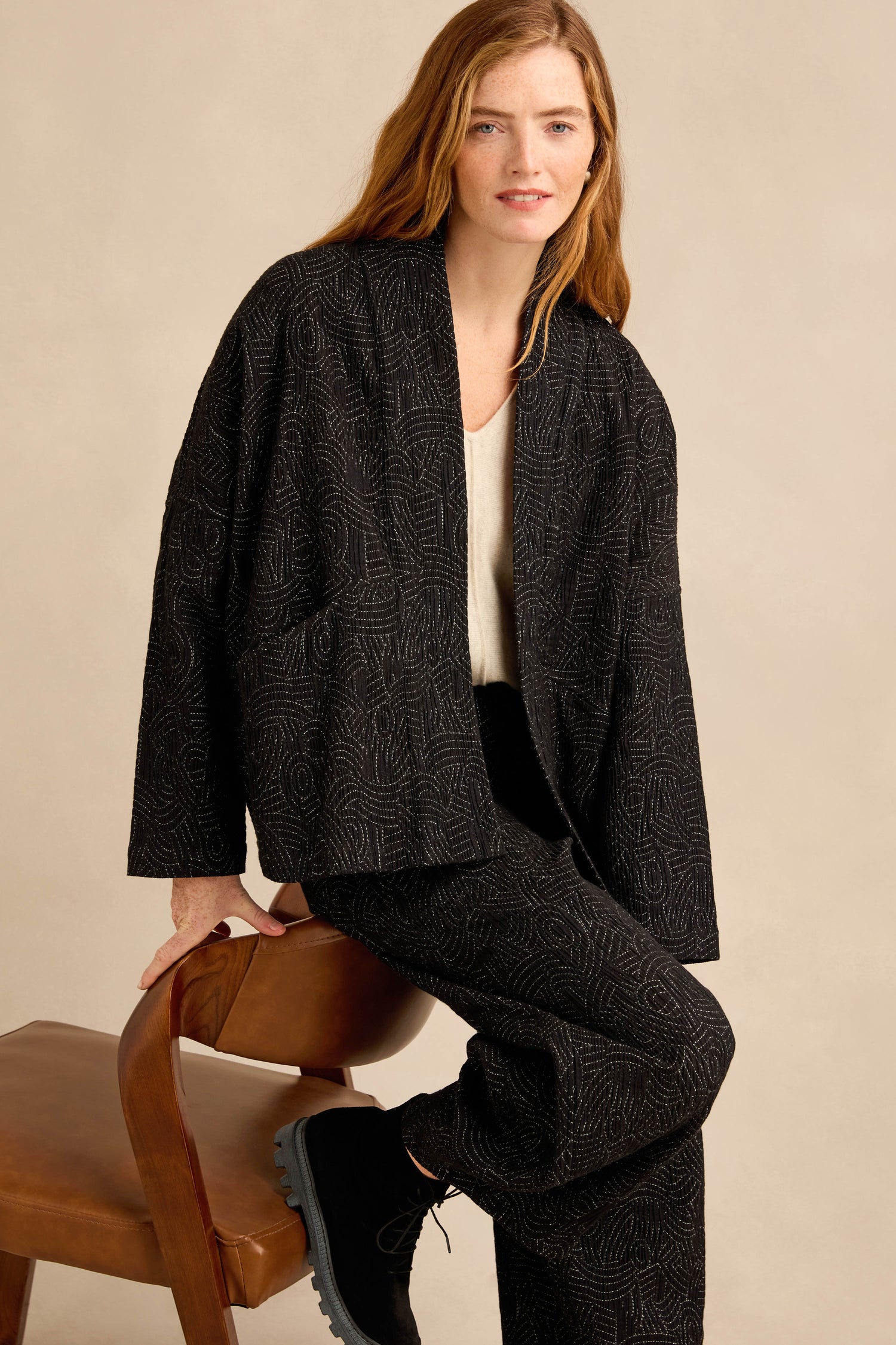 A person with long hair sits on a wooden chair against a plain beige background, wearing the Stitched Jacquard Jacket and matching pants, paired with a light-colored top and black boots. The jacket’s boxy silhouette lends modern structure to the ensemble.