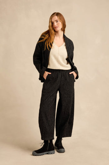 A woman stands with her hands in her pockets, wearing a cream V-neck top, the Stitched Jacquard Jacket with a boxy silhouette, matching wide-leg pants, and black boots against a beige background.
