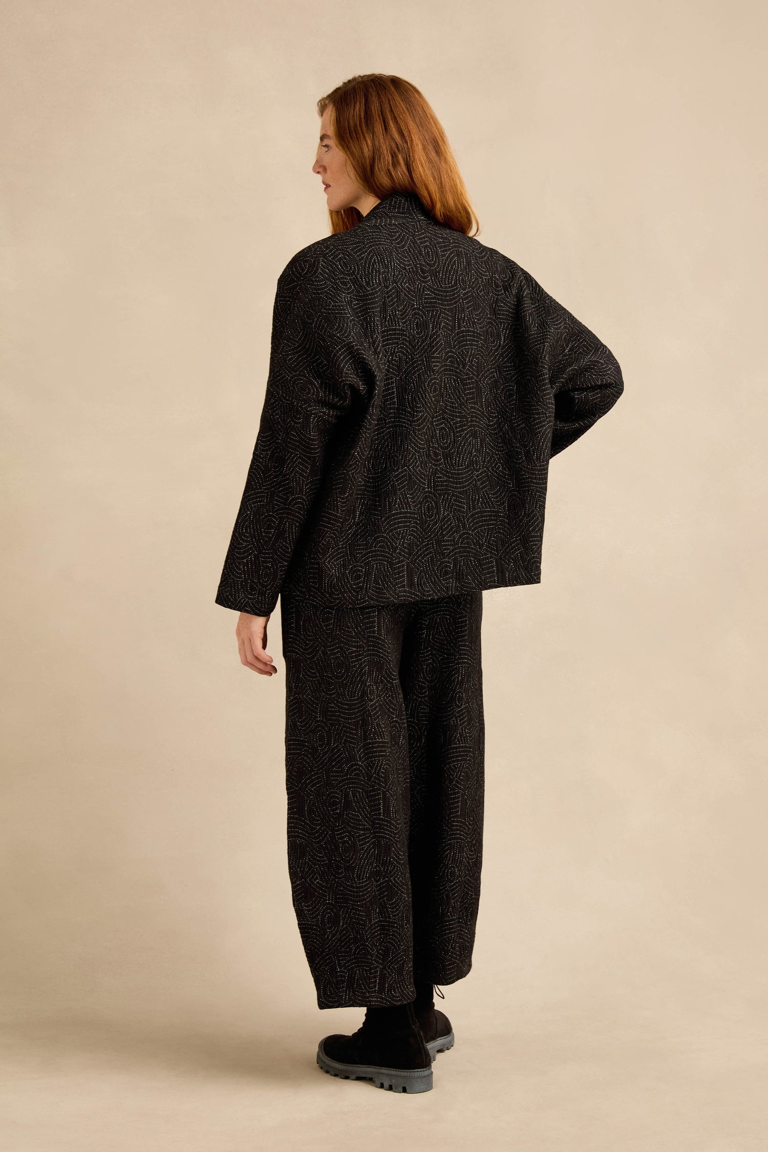 A person with long auburn hair, wearing the Stitched Jacquard Jacket in pure black cotton with a subtle quilted pattern and paired with black shoes, stands facing away against a beige background.