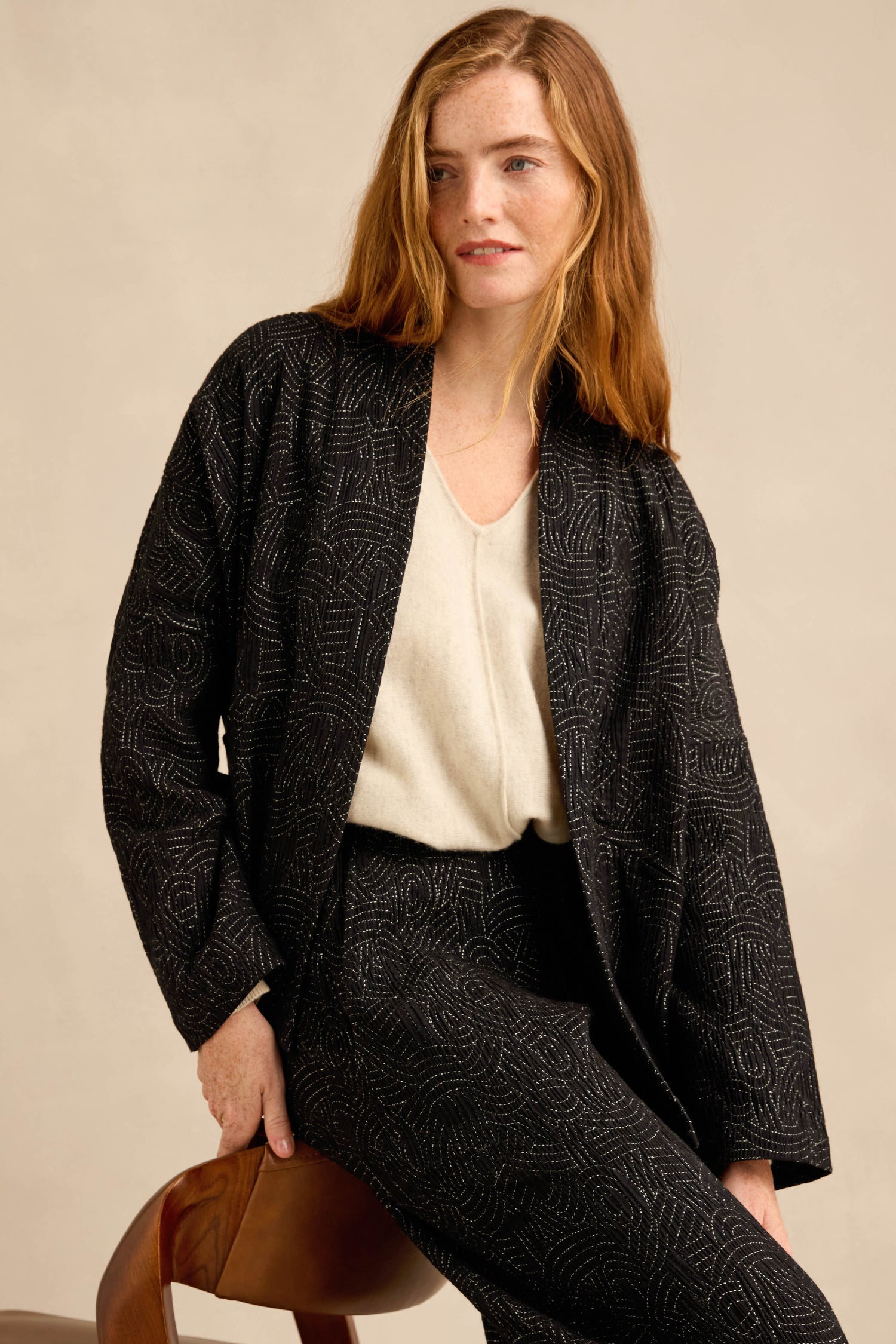 A woman with long red hair wears the Stitched Jacquard Jacket in a boxy silhouette over a cream top, seated on a brown chair against a plain beige background.
