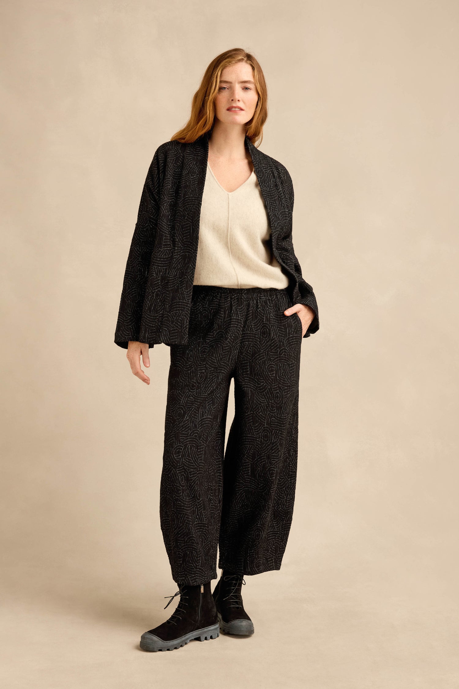 A woman stands against a beige background wearing the Stitched Jacquard Jacket in a boxy silhouette, paired with a cream V-neck top and black boots, with one hand in her pocket.