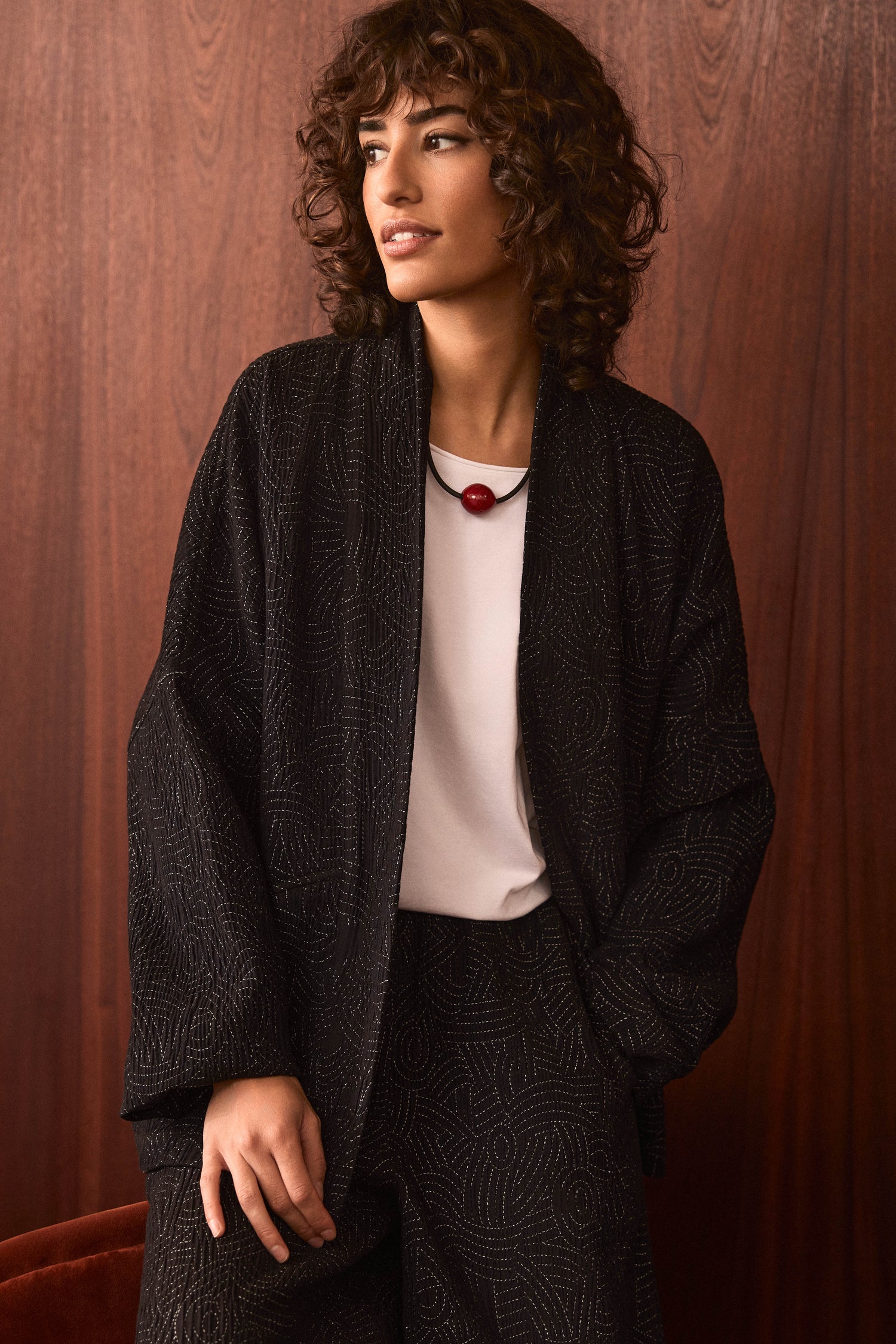 A person with curly hair is wearing the Stitched Jacquard Jacket, which features a quilted pattern, paired with a white shirt and a necklace adorned with a red bead, standing in front of a wooden wall.