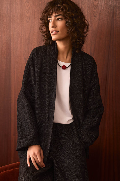 A person with curly hair is wearing the Stitched Jacquard Jacket, which features a quilted pattern, paired with a white shirt and a necklace adorned with a red bead, standing in front of a wooden wall.