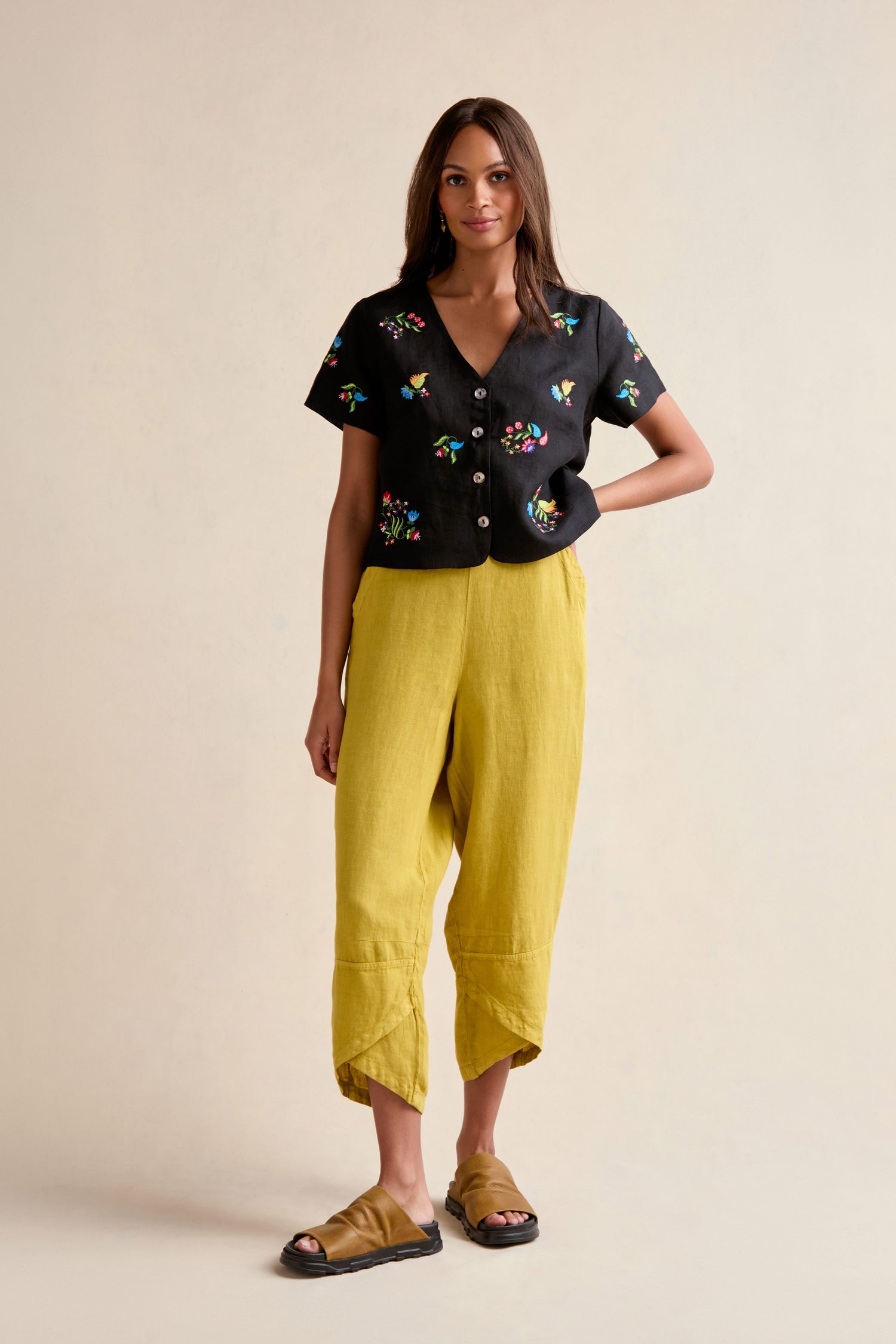 Woman against a plain background, wearing a Vintage Floral Embroidered Jacket, pure linen loose yellow pants, and tan slide sandals.